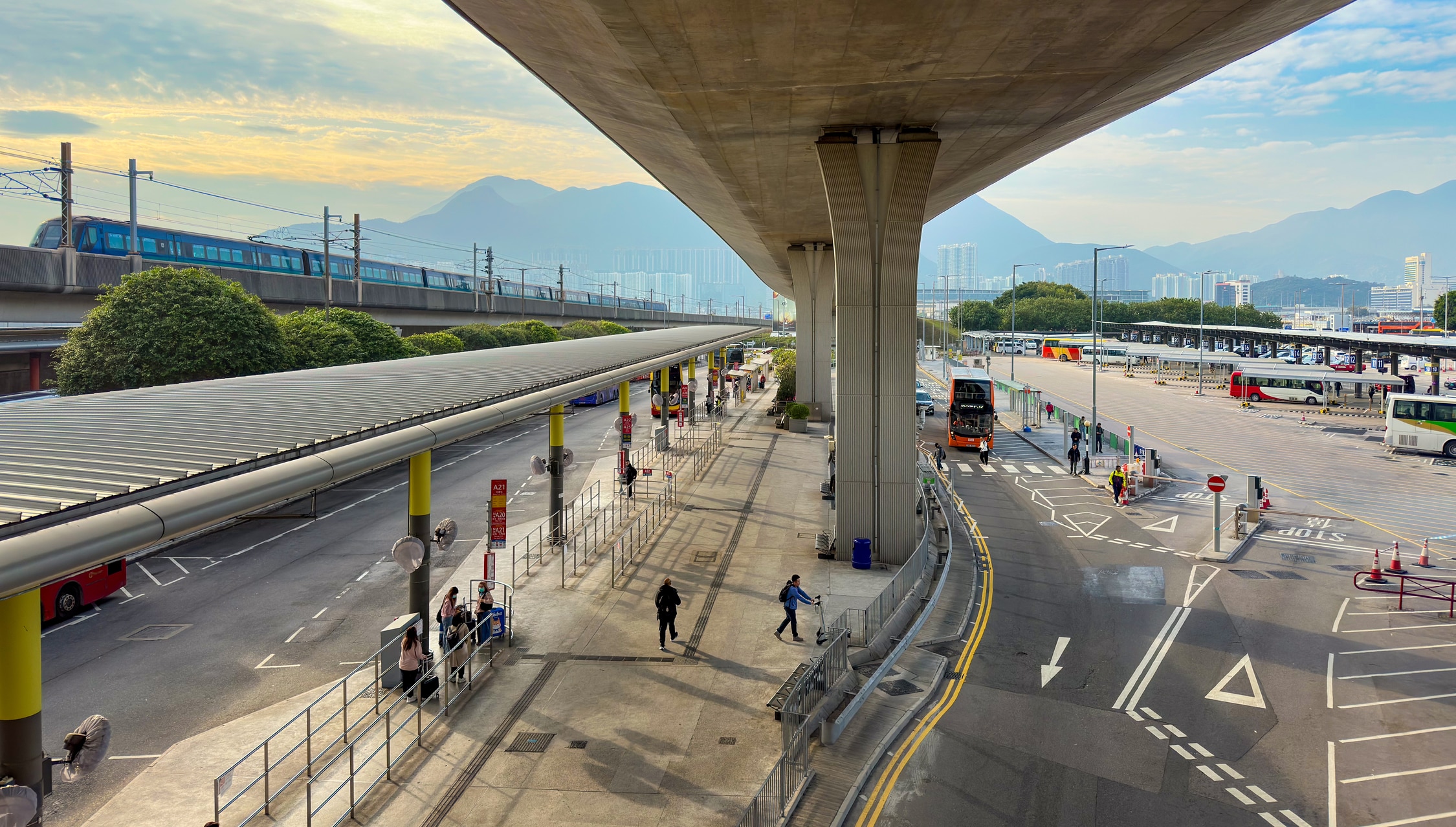 Blick unter einer Brücke entlang der Fahrbahn. Links ist die Brücke mit einem Zug drauf. unten ist eine große Bushaltestelle zu sehen. Im Hintergrund hohe Berge.