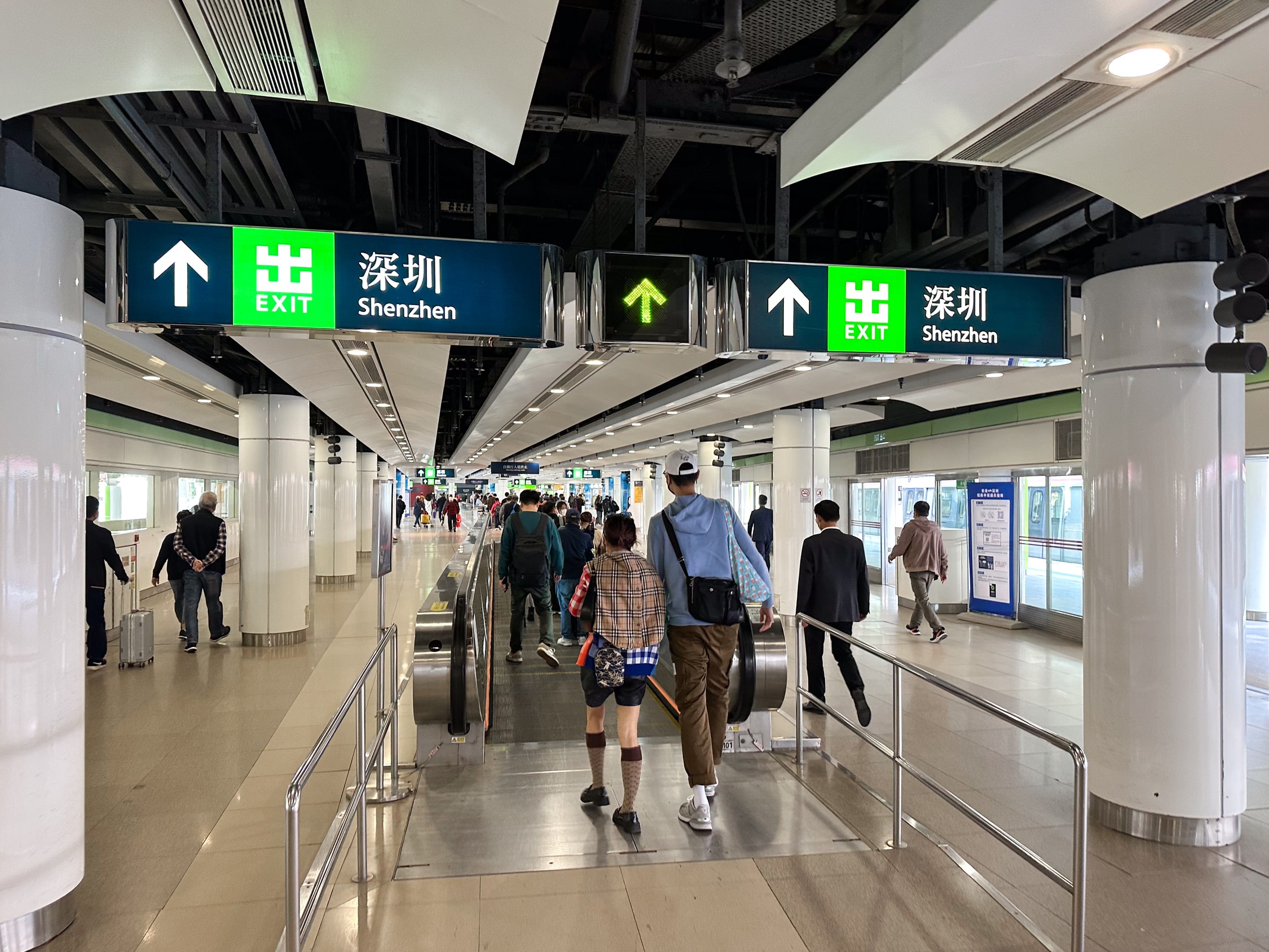 Ein langer Gang mit Laufbändern. Darüber Schilder auf denen Exit und Shenzhen steht.
