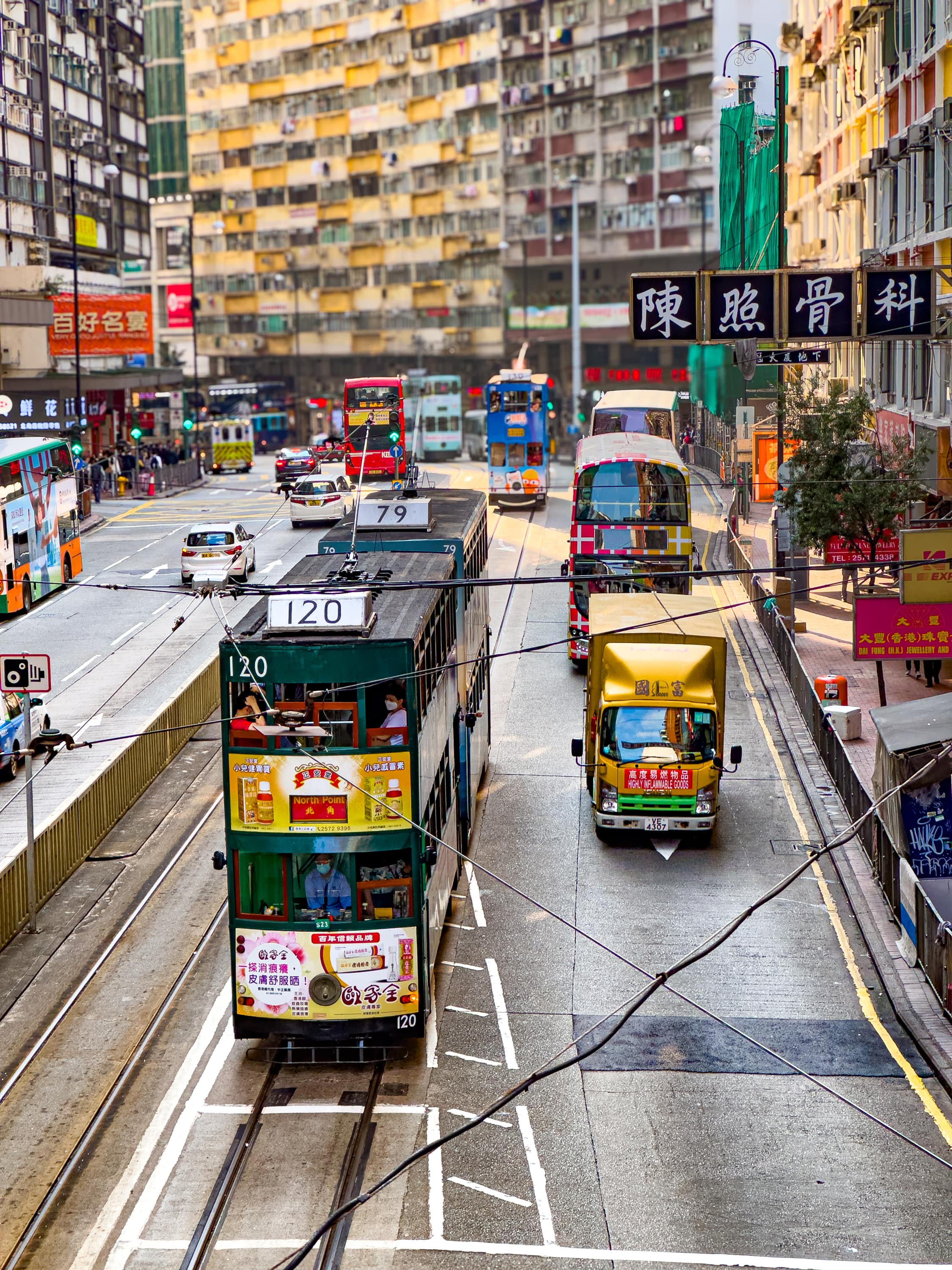 Eine Straße mit Hochäusern im Hintergrund. Über die Straße ragen Schilder mit chinesischen Schriftzeichen. Auf der Straße einige Trams, Doppelstockbusse, LKWs und Autos