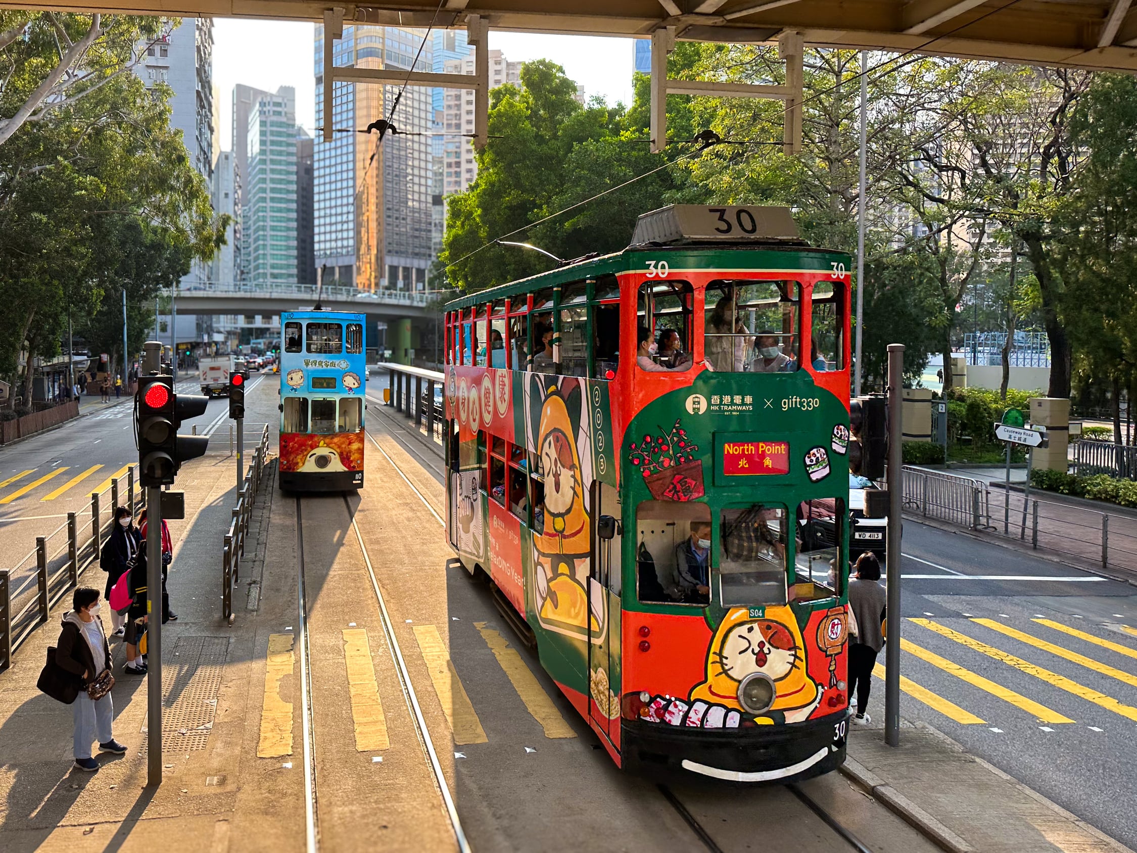 Zwei Doppelstockstraßenbahnen begegnen sich. Sie werfen Schatten von der Tiefstehenden Sonne. Beide sind mit Cartoontieren bemalt. Die eine mit einer Katze auf grünen und roten Hintergrund. Die andere mit Hühnchen auf blauem Hintergrund