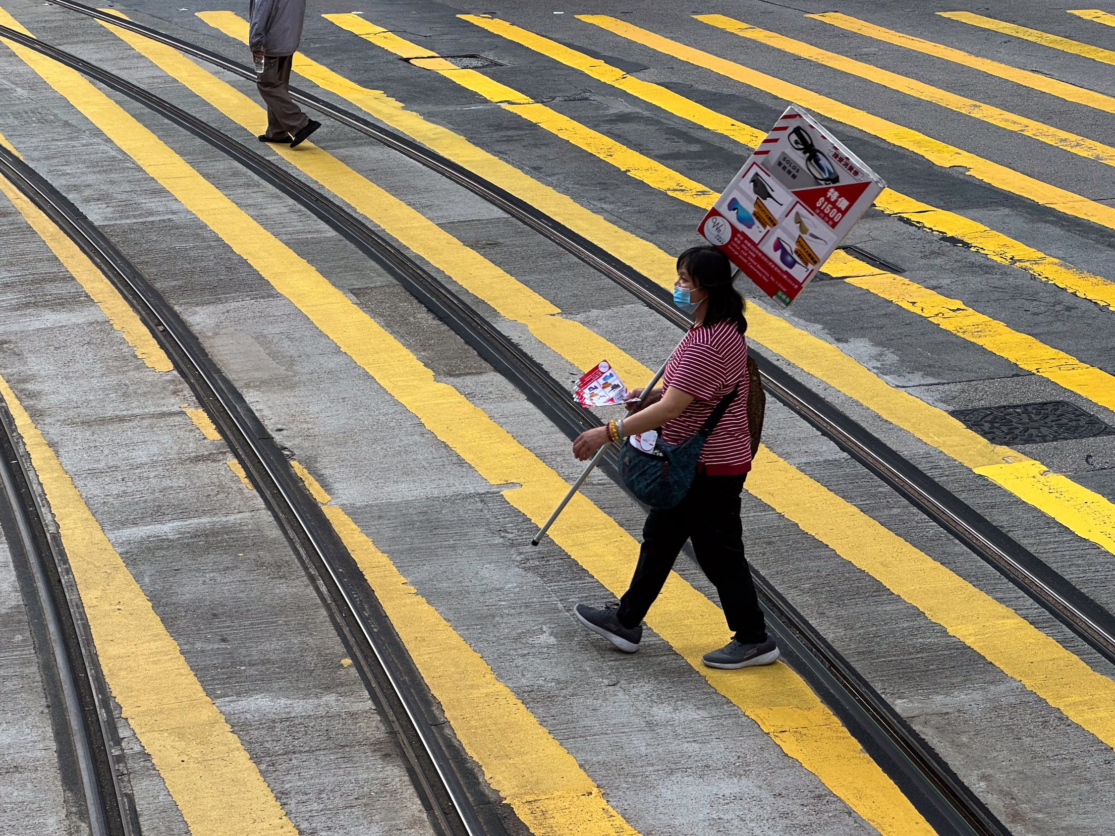Eine Frau läuft über einen rieseigen Zebrastreifen, der über die Straßenbahnschienen führt. Sie trägt ein Werbeschild für ein Brillengeschäft und hält Flyer für dieses in der Hand.