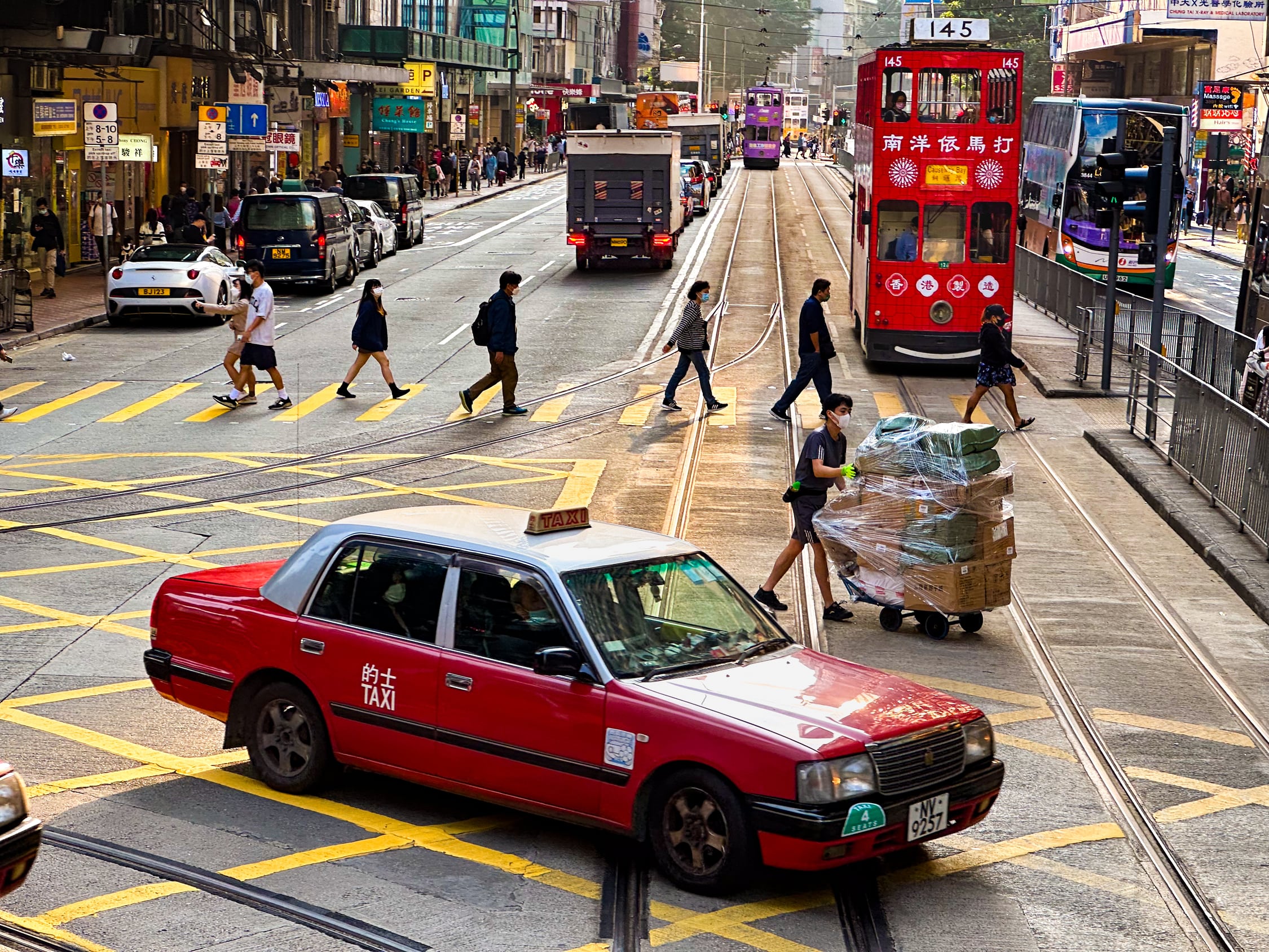 Eine Kreuzung mit abzweigenden Straßenbahngleisen in Gegensonne. Gegenüber eine weitere Straßenbahn. Ein rotes Taxi kreuzt den Weg der Bahnen. Über einen Zebrastreifen laufen Menschen. Ein Mensch mit Kisten auf einem großen Handwagen Kreuzt die Straßenbahntrasse. Auf den Gehwegen laufen viele Menschen. Auf der Rechten Straßenseite kommt ein Doppelstockbus entgegen, auf der linken fahren Autos und ein LKW. In der Ferne weitere Doppelstocktrams