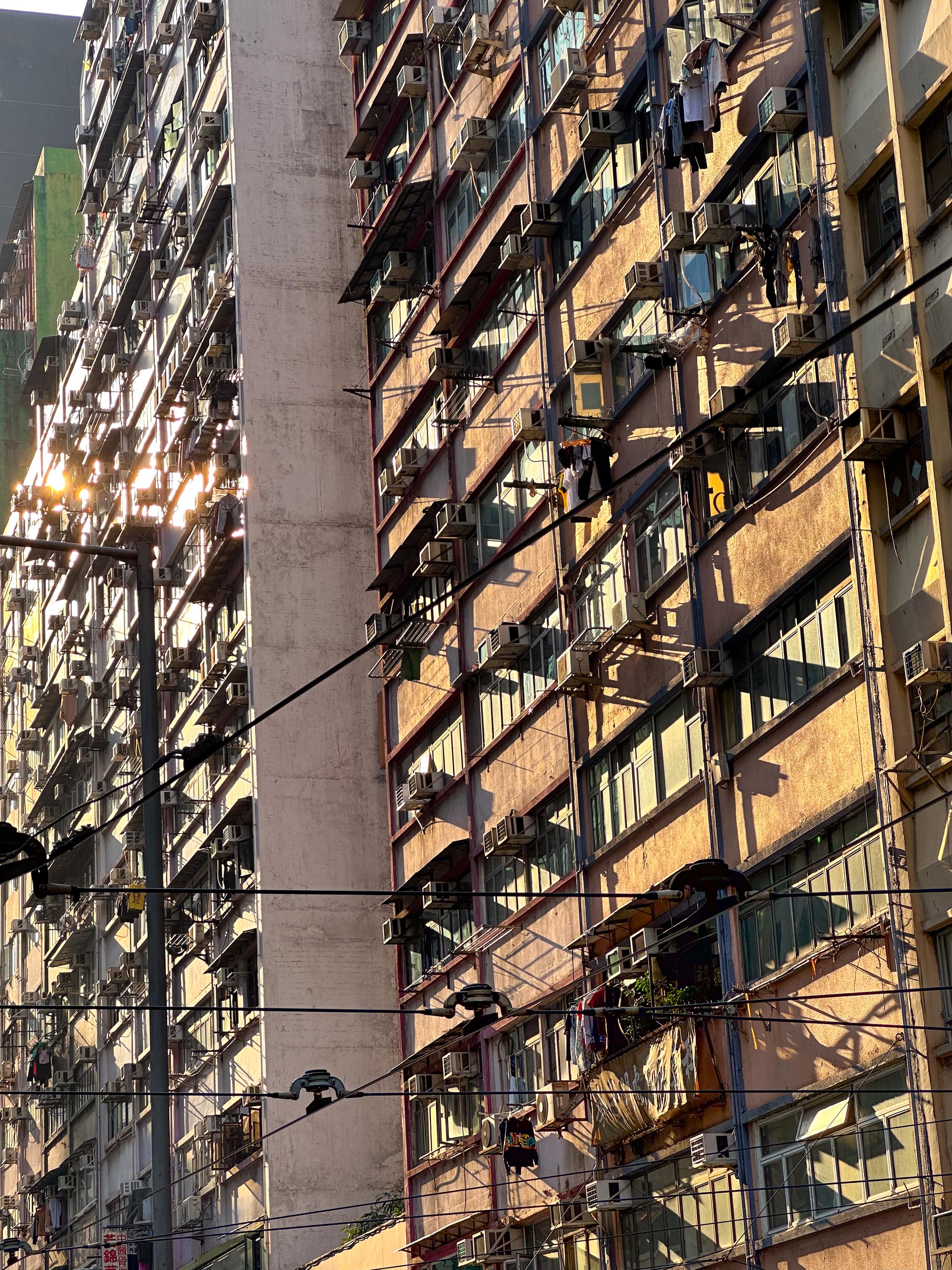Im Steilen Winkel aufgenommende Fassade der Wohnhochhäuser. In einigen Fenstern spiegelt sich die Sonne. Aus vielen Fenstern wird Wäsche gehängt. Viele Außenklimanlagen, oder Balkonartige bauten. Es ist viel Oberleitungskonstruktion zu sehen
