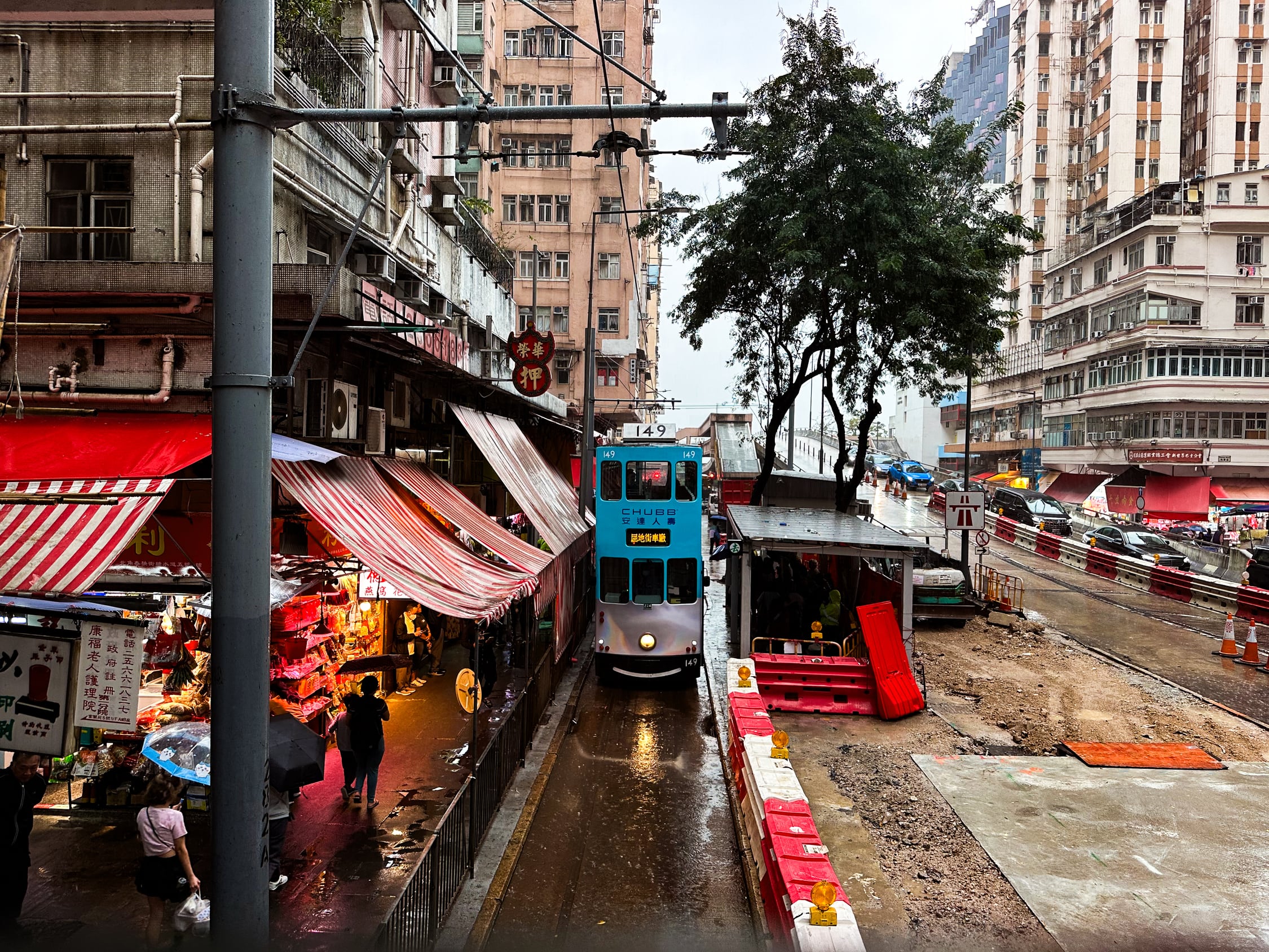 Blick vom oberen Deck einer Tram auf einen eingleisigen Streckenabschnitt. Es ist regnerisch und Menschen trsgen regenschirme und schützen sich unter Markiesen der Gebäude.