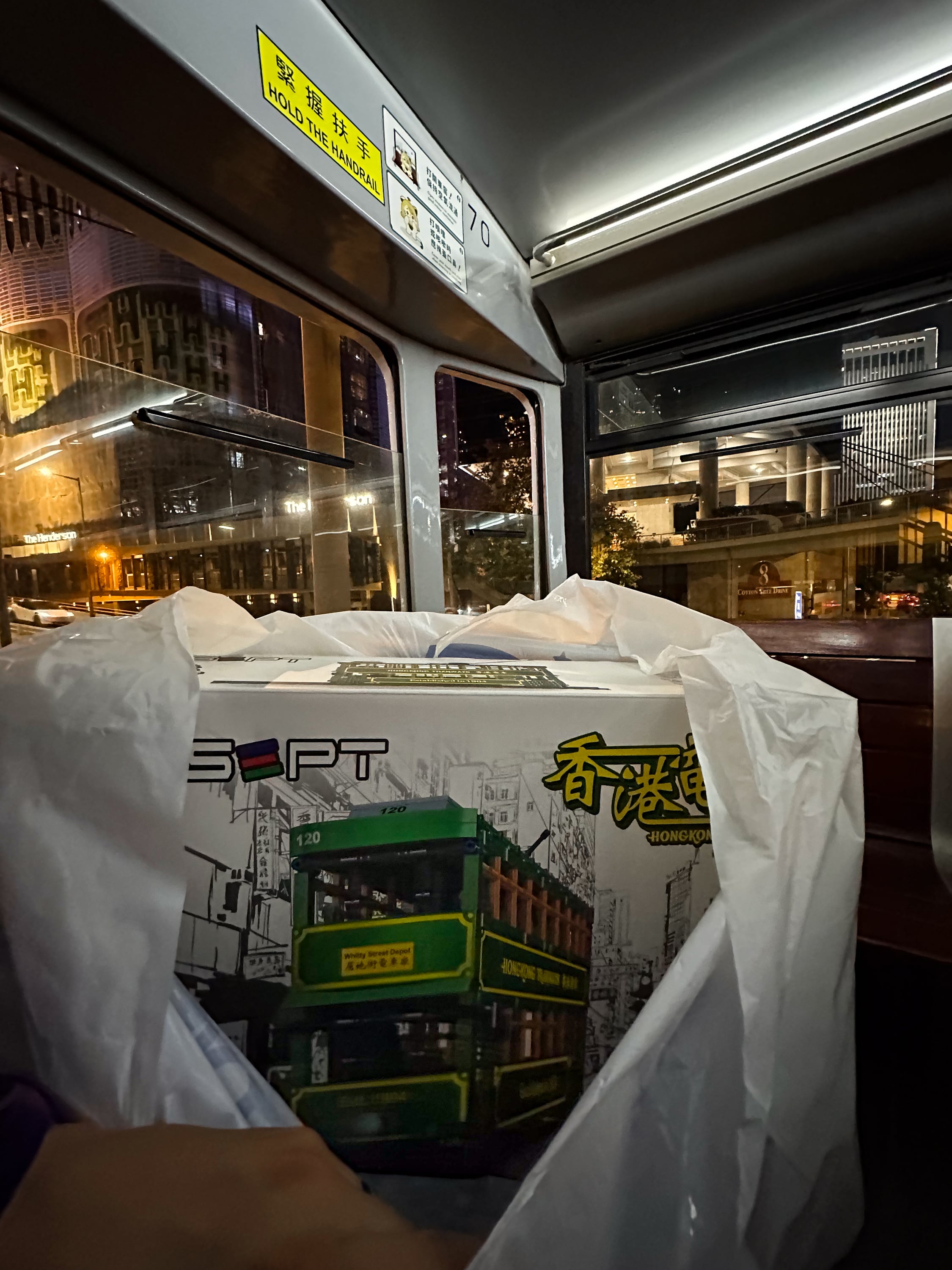 Ein Beutel, der so aufgezogen ist, dass man sieht, dass sich darin ein riesiger Karton befindet, auf dem eine Lego-Doppelstocktram abgebildet ist. Im Hintergrund ist das Innere des oberen Decks einer Doppelstocktram zu sehen