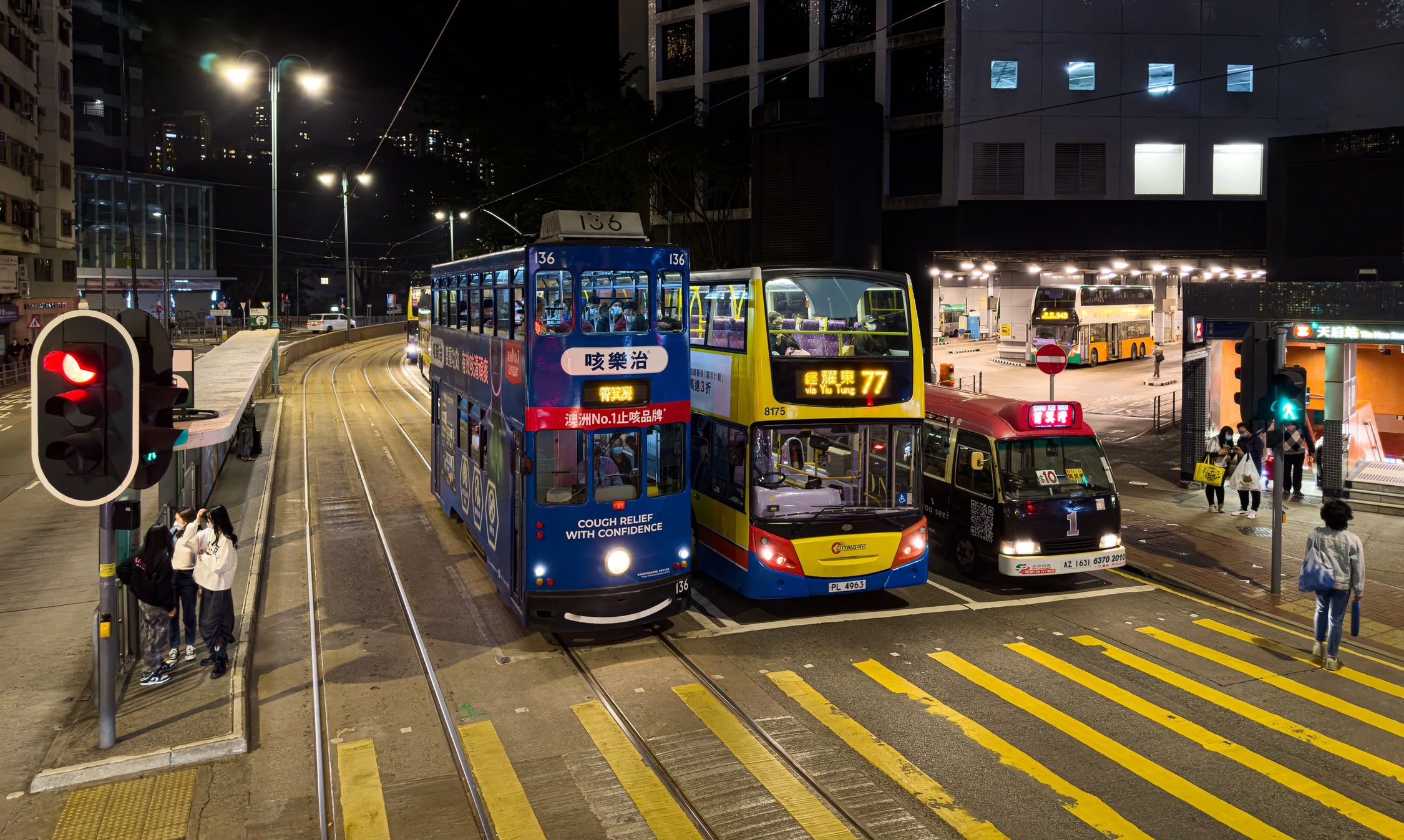 Eine Doppelstocktram, ein Doppelstockbus und ein Minibus stehen nebeneinander an einer Ampel. Dahinter die Einfahrt zu einem Busbahnhof in einem Gebäude. An der Seite der eingang zu einem U-Bahnhof.