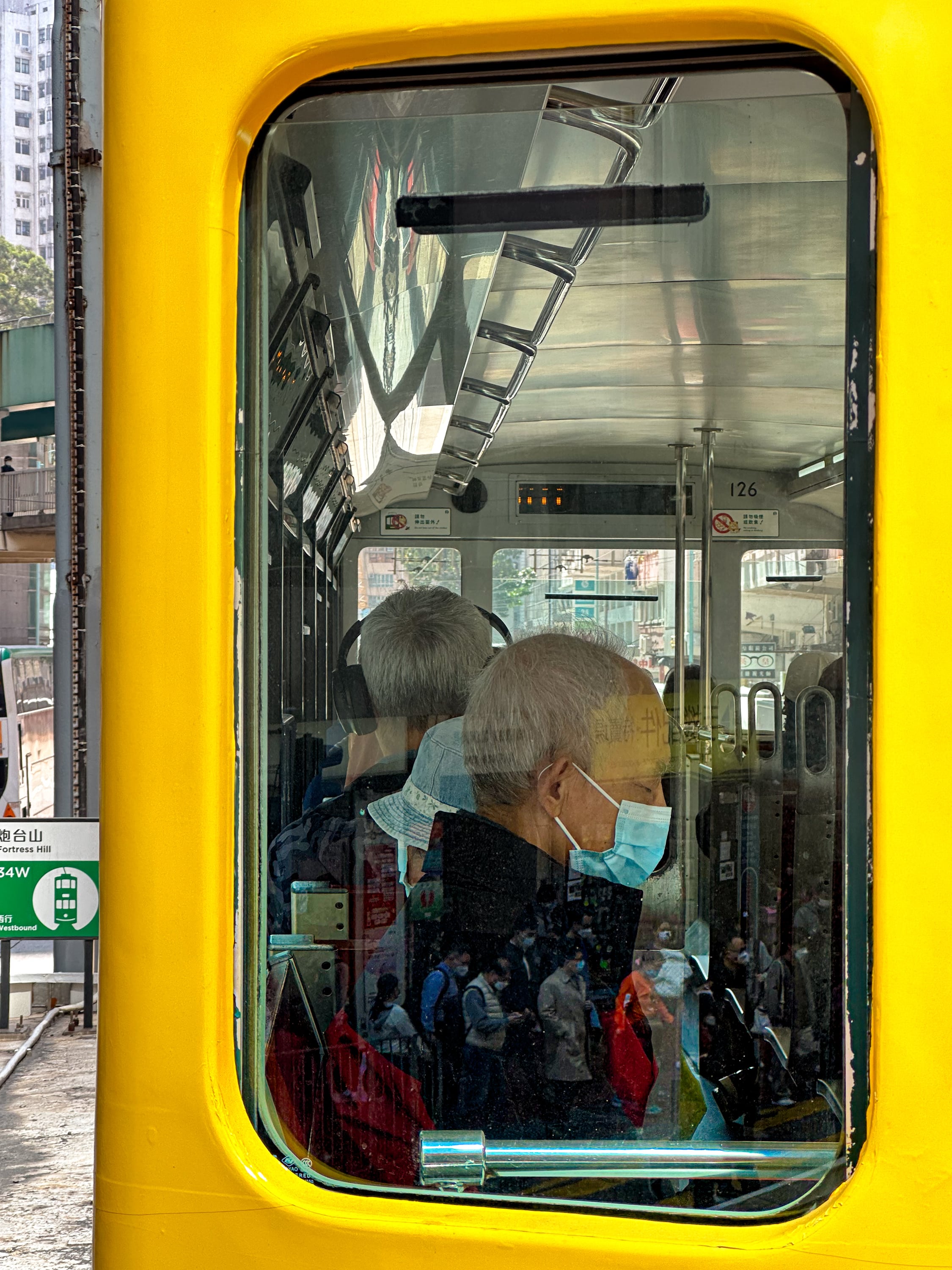 In das linke Fenster des oberen Decks der vorrausfahrenden Tram fotografiert. Dahinter sitzt ein Mann mit Maske. In dem Fenster Spiegeln sich wartende Menschen auf der Straße. Links neben der Bahn ist das Schild der Haltestelle Fortress Hill zu erkennen