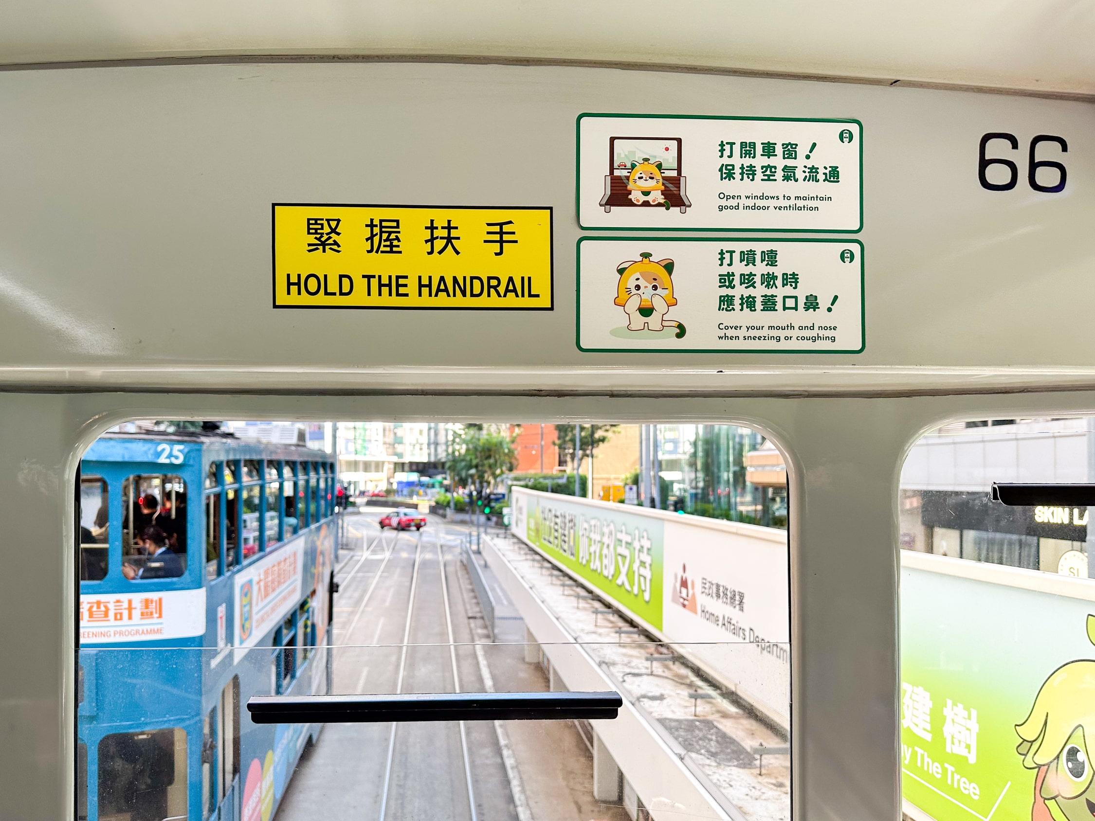 Schilder über dem vorderen Fenster. 'Hold the Handrail', 'Open the window to maintain doof indoor ventialation' daneben eine cartoon Katze, die auf eine Bank sietzt. 'Cover you mouth and nose, when sneezing' daneben die Cartoon Katze die sich die Pfoten vor den Mund hält. Durch das halb geöffnete Fenster ist eine Doppelstrock-Tram auf der Gegenspur zu sehen