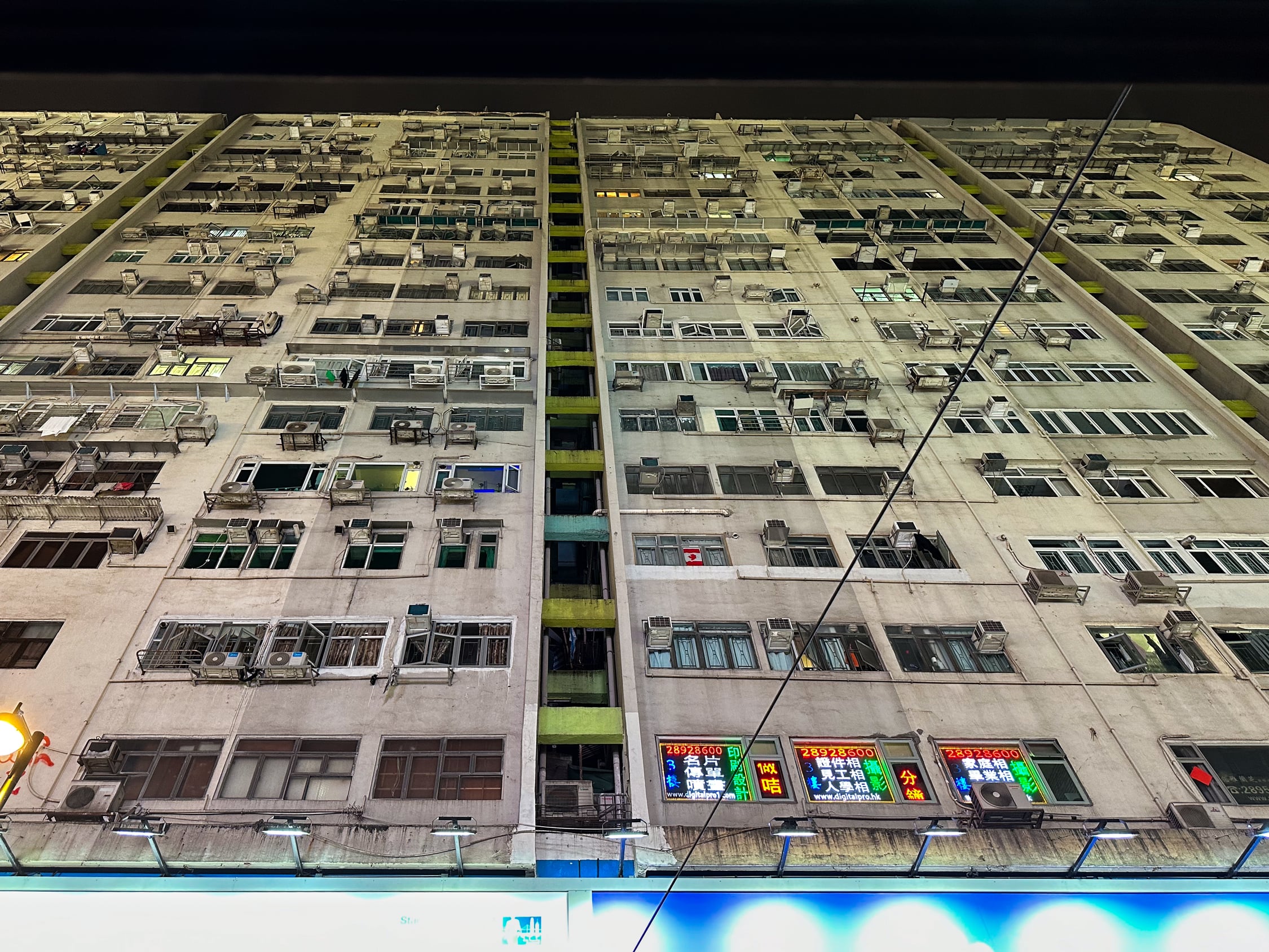 Aus dem Seitenfenster der Tram nach oben fotografiertes Wohnhaus mit etwa 20 Stockwerken. Es ist nach und einige Fenster erleuchet in den unteren Stockwerken sind Bunte Werbetafeln aus durch die Fenster zu sehen.