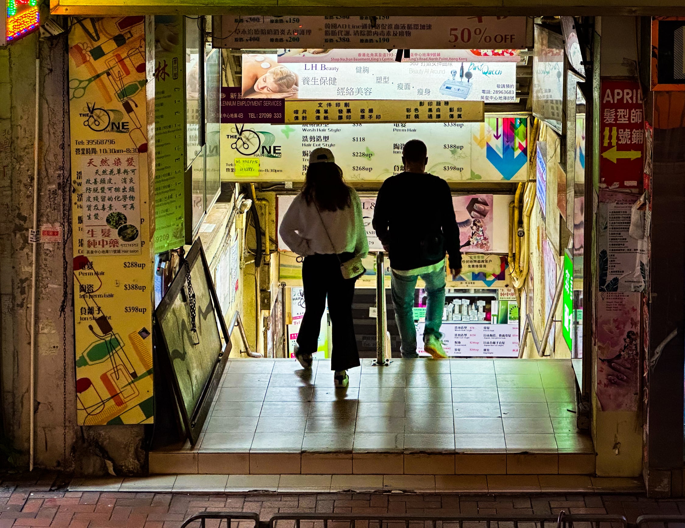 Blick in einen Geschäftseingang, der direkt mit einer Treppe nach unten Führt. Da aus dem Oberen Deck der Tram fotografiert wurde, kann man etwas weiter die Treppe nach unten sehen. Überall an der Deckenschränge sind Schilder für die unten Liegenden Geschäfte angebracht. Zwei menschen gehen die Treppe nach unten.