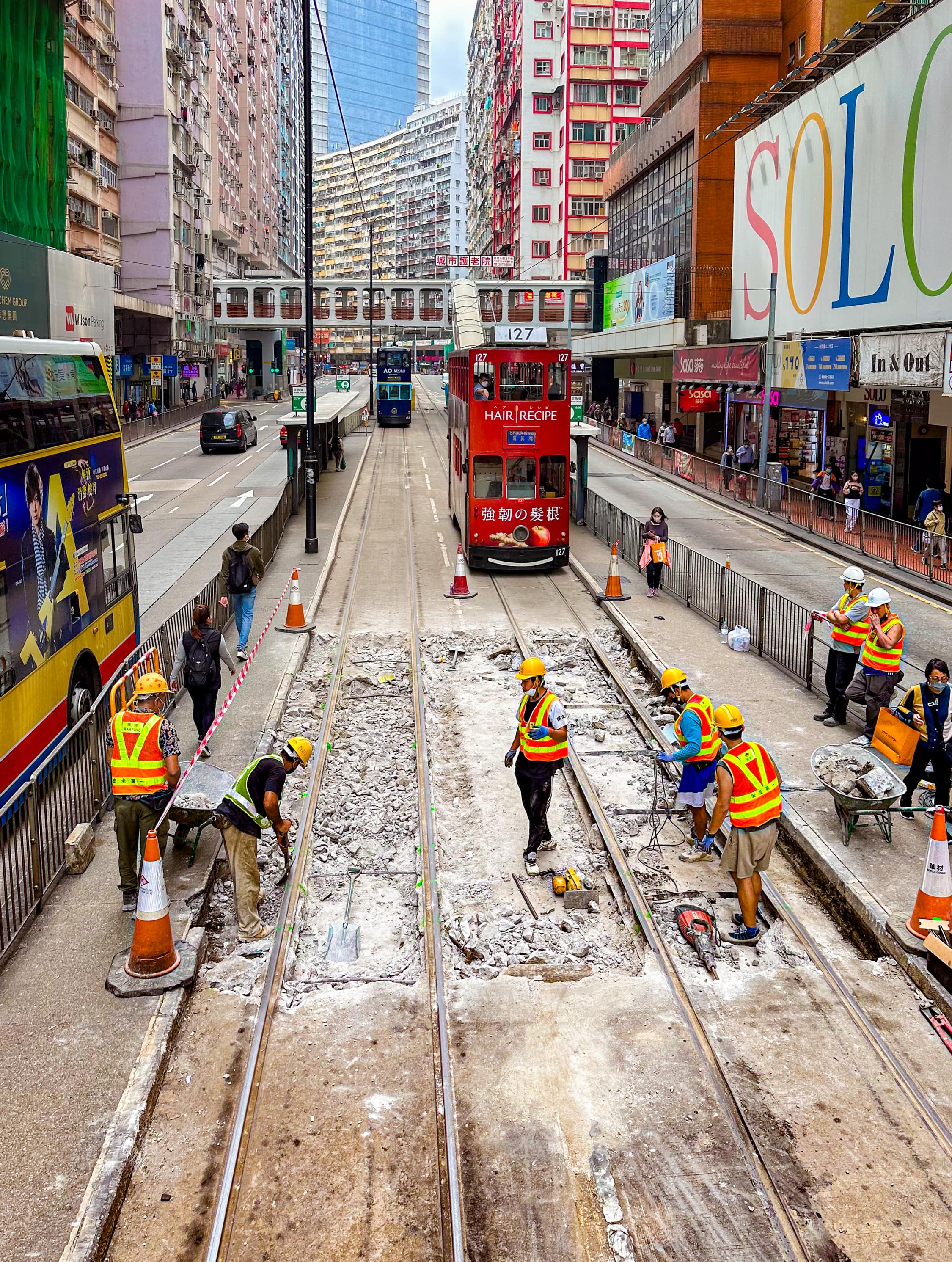 Vor der Straßenbahn sind etwa 5 Meter Straße über die komplette breite der beiden Gleise aufgebrochen. 7 Bauarbeiter arbeiten mit Schaufel, Presslufthammer und Schubkarre an den Gleisen. Auf der gegenüberliegenden Seite wartet eine Tram, aber auf der gleichen Spur ist auch eine Tram zu sehen, die wenige Sekunden vorher die Baustelle passiert hat.