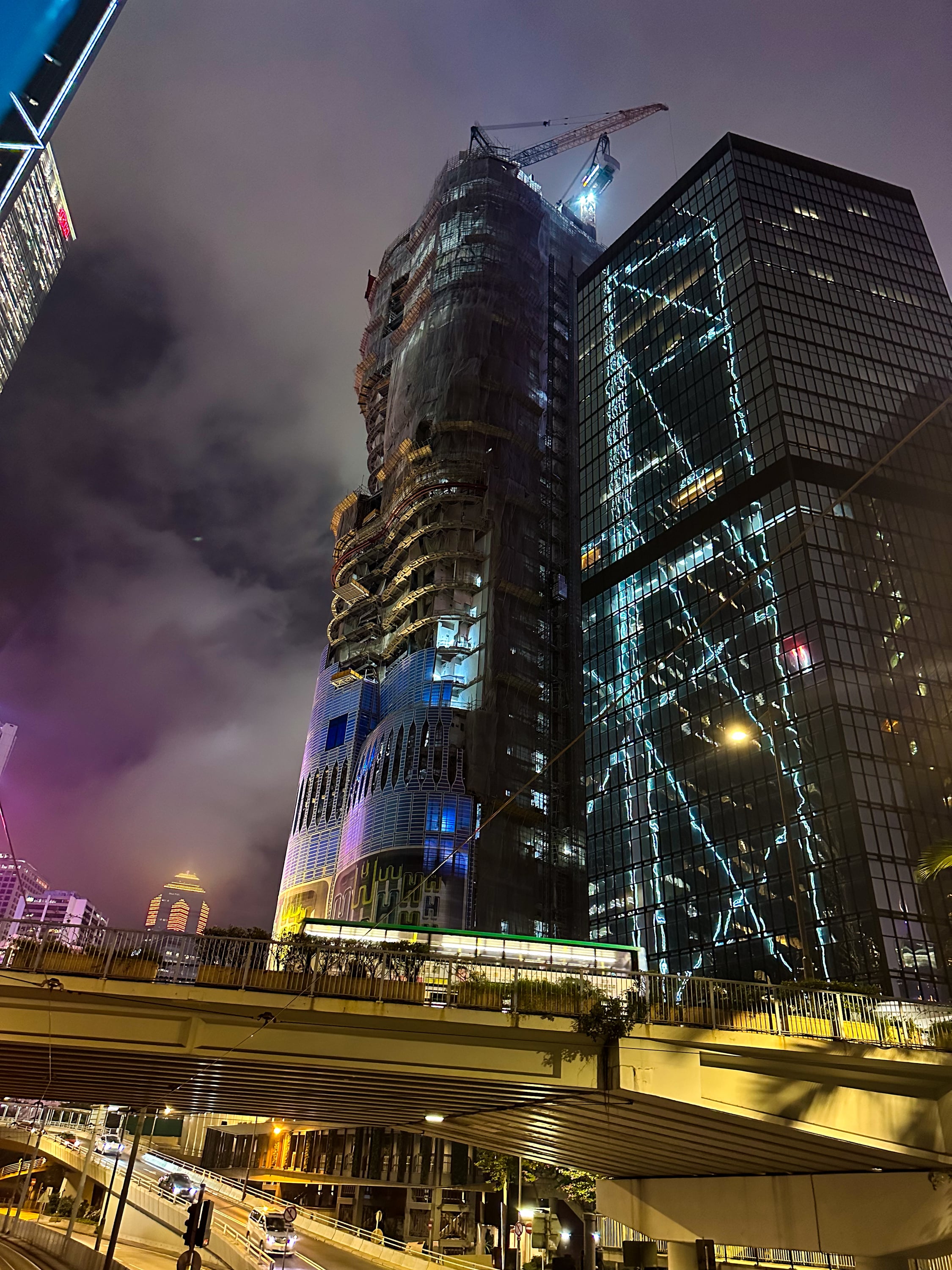 Nächtlich. Blick nach oben auf ein im bau befindliches Büro-Hochhaus. In dem Büro-Hochhaus daneben spiegeln sich die Lichteffekte des Bank of China Towers. Es ist leicht bewölkt oder gar nebelig, so dass die Lichter der Werbung oder der gerade stattfinden Lichtshow den Himmel lila verfärben. Weiter unten ist die Straße und eine Brücke zu sehen über die ein Doppelstockbus fährt. Im Hintergrund ein weitreres bunt beleuchtetes Hochhaus.
