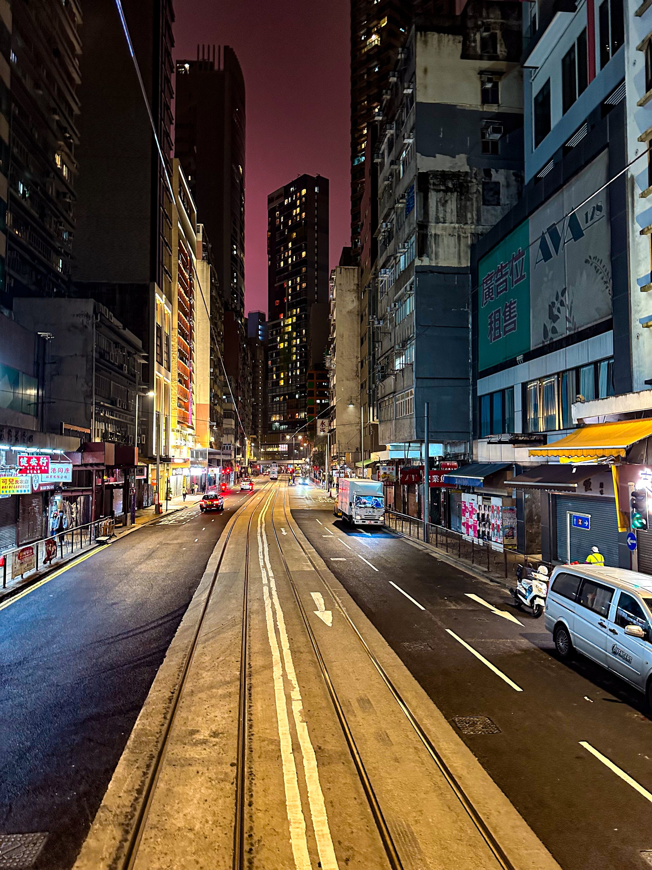 Nächtliche Straße, von oben aus der Tram fotografiert. relativ leer. Am Ende Straße ein großes Hochaus. der Himmel ist lila, wahrscheinlich von der vielen LED-Werbung an den Hochhäusern in der Stadt