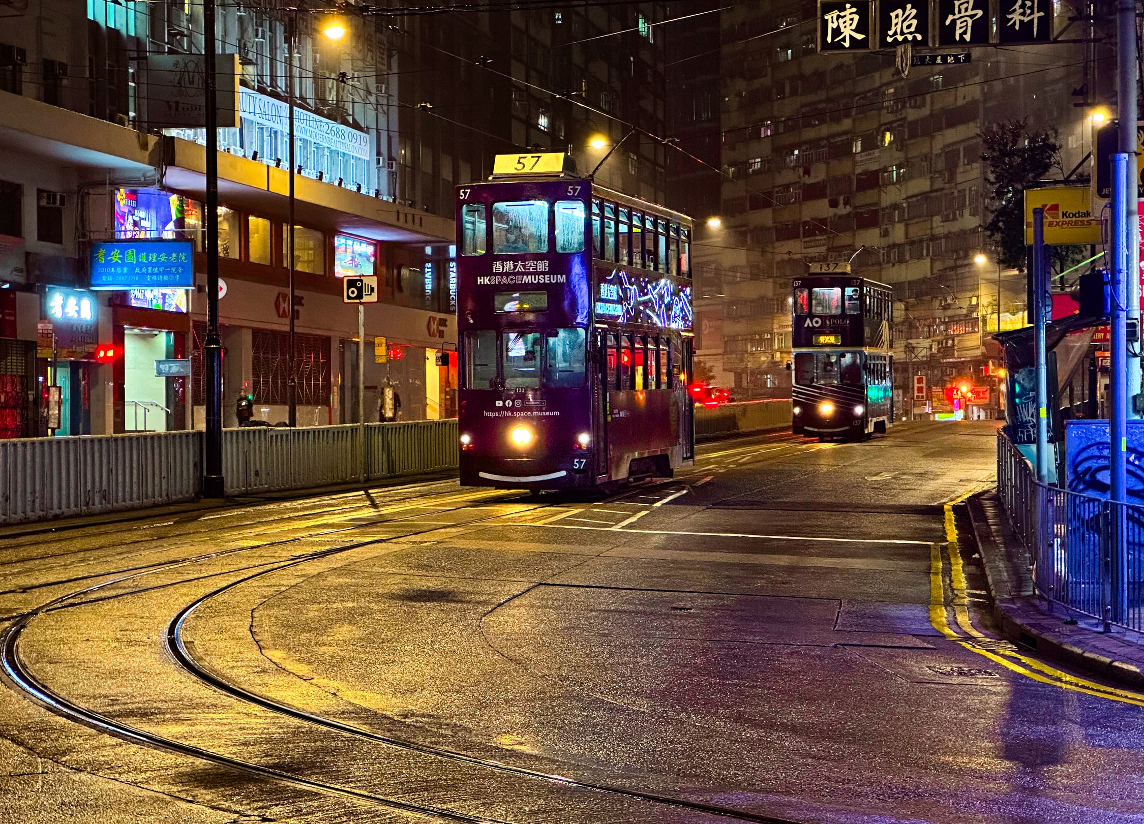 Zwei Trams fahren nachts durch die Straße. Der Boden ist nass und spiegelt das Licht. Eine Tram macht Werbung für das Hong Kong Space Mueseum. Der Schriftzug des Museums und ein Weltraummuster an der Seite der Tram leuchten.