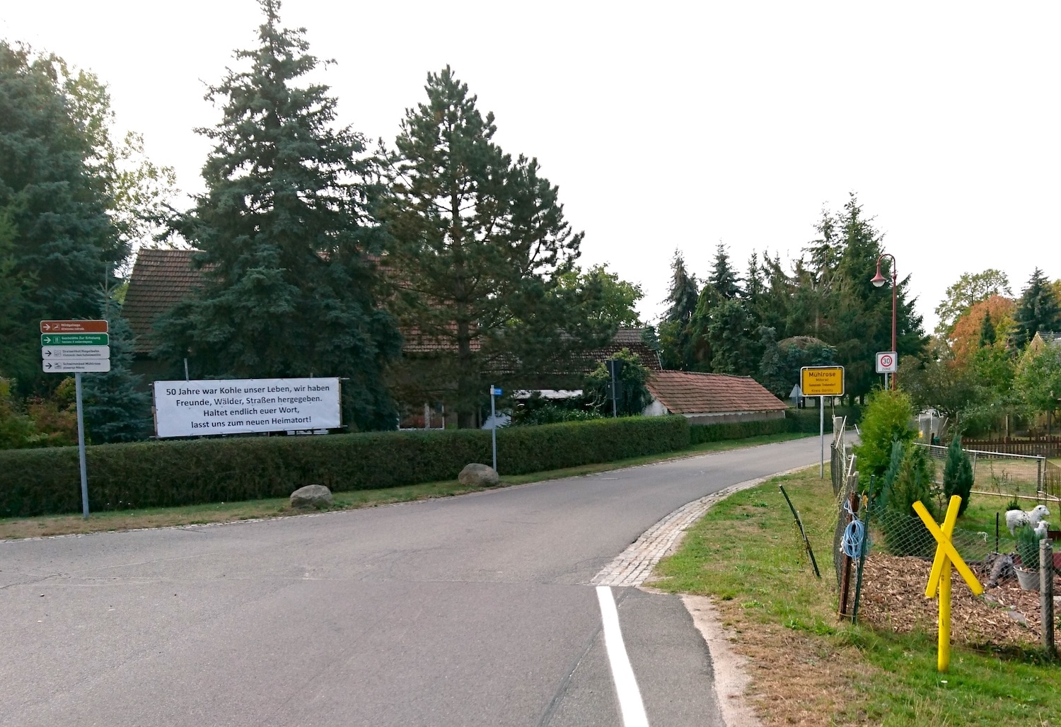 Ortseingang von Mühlrose mit einem Protestschild für die Umsiedelung aber auch einem gelben Kreuz für die Erhaltung des Ortes