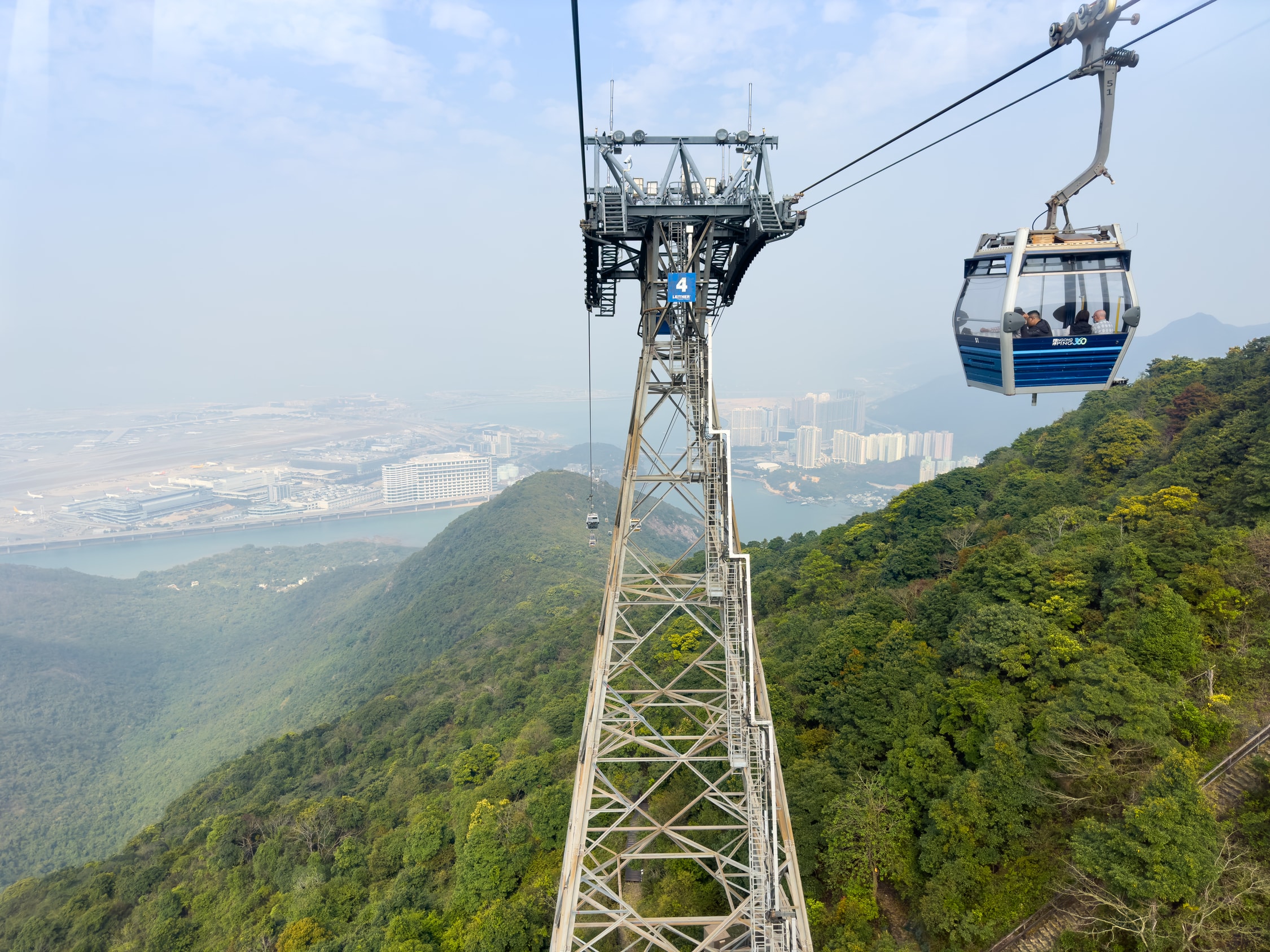 Ein Stahlgittermast auf einem Berg. In Höhe der Seilbahnführung sind Scheinwerfer, Mobilfunkantenen und Überwachungskameras angebracht. Im Hintergrund sieht man den Berg hinunter. Dort sieht man im Nebel eine Insel mit Gebäuden drauf.