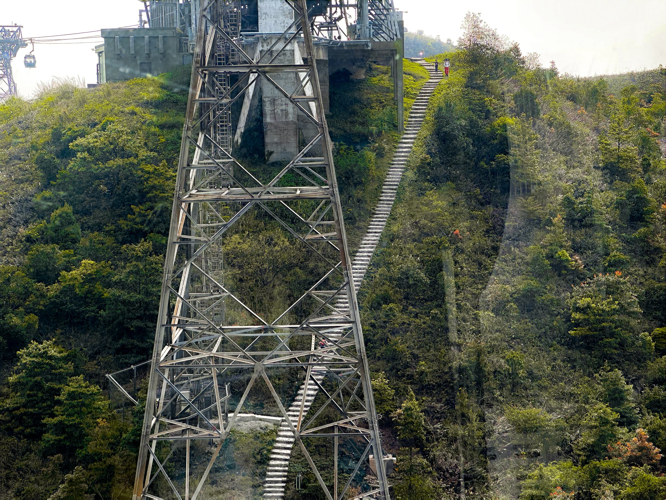 Blick durch einen Stahlgittermast. Auf die dahinter liegende Bergspitze, auf der sich eine Winkelstation befindet führt eine sehr lange steinernde Treppe. Auf dieser sind ein paar Wandererinnen zu sehen.