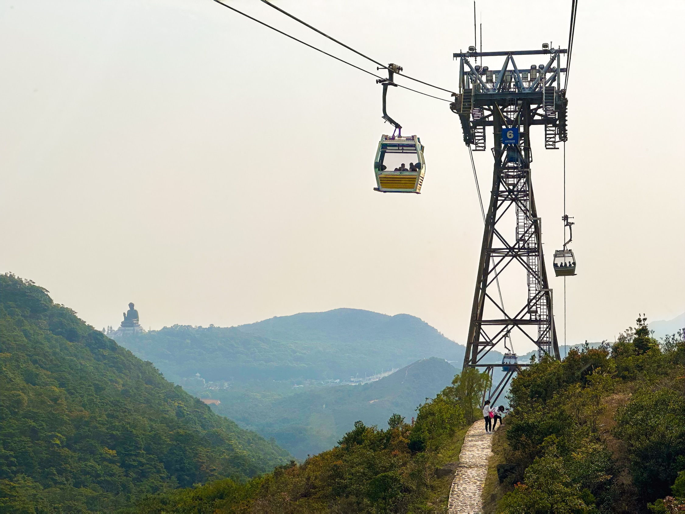 Ein Weiterer Mast durch den der Weg führt. Am Rande des Weges rasten zwei Wandererinnen. Im Hintergrund ist der Berg mit dem Buddha zu sehen.