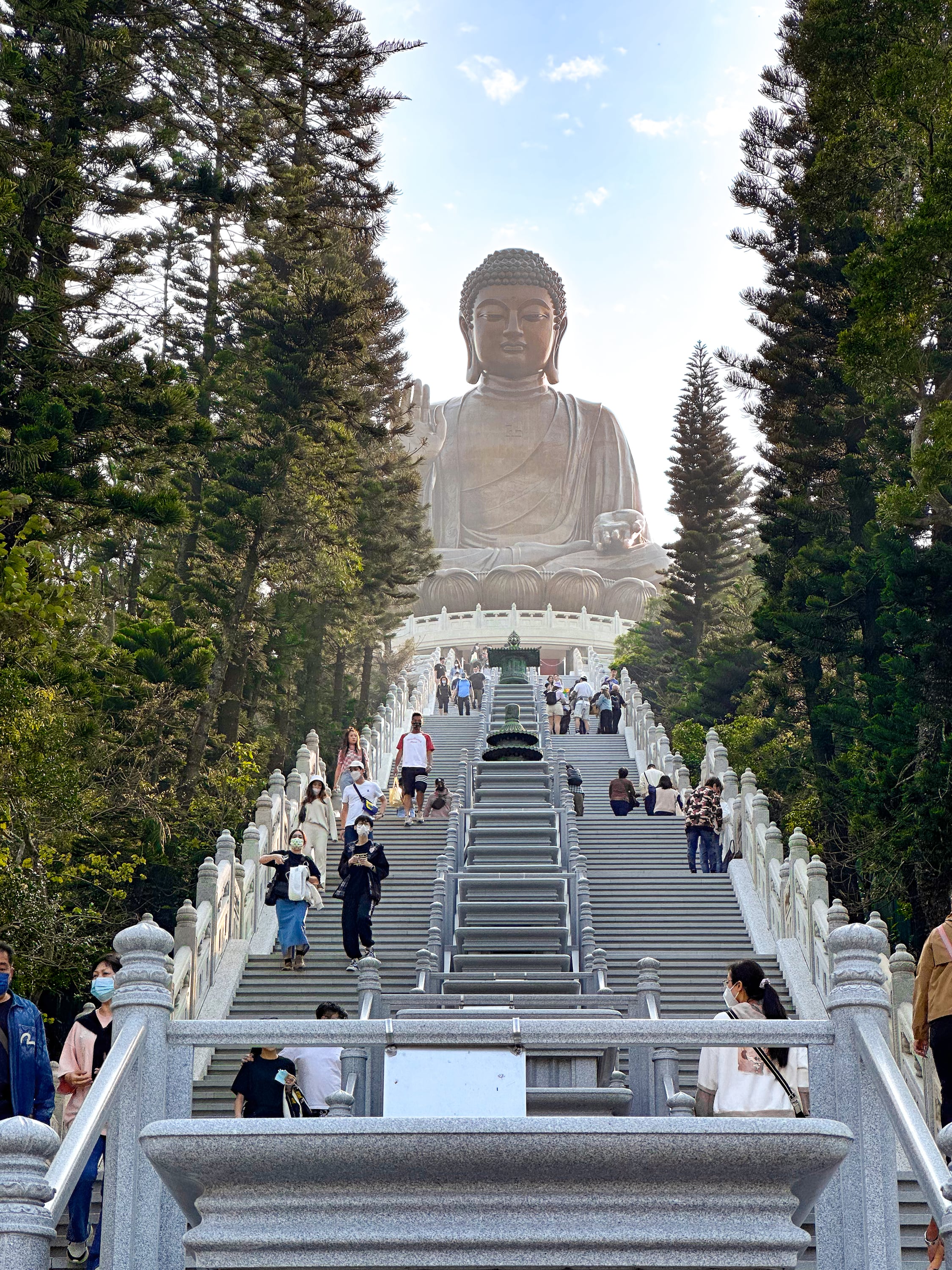 Der Sitzende Buddah auf dem Berg. Vor ihm eine Treppe die den Berg zu ihm hoch führt. Auf der Treppe viele Menschen
