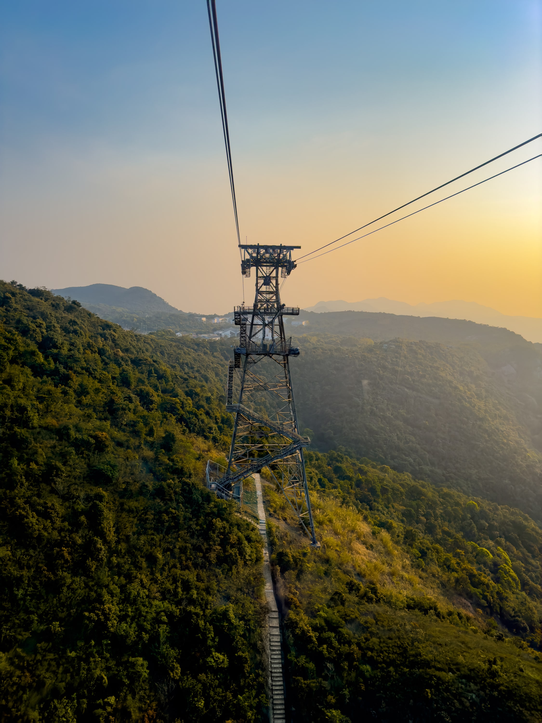 Strahlgittermast für die Seilbahn an einem Berghang. Unter dem Mast führt ein Weg hindurch. Eine Treppe führt den Mast hinauf. Im Hintegrund Berge und Sonnenuntergang.